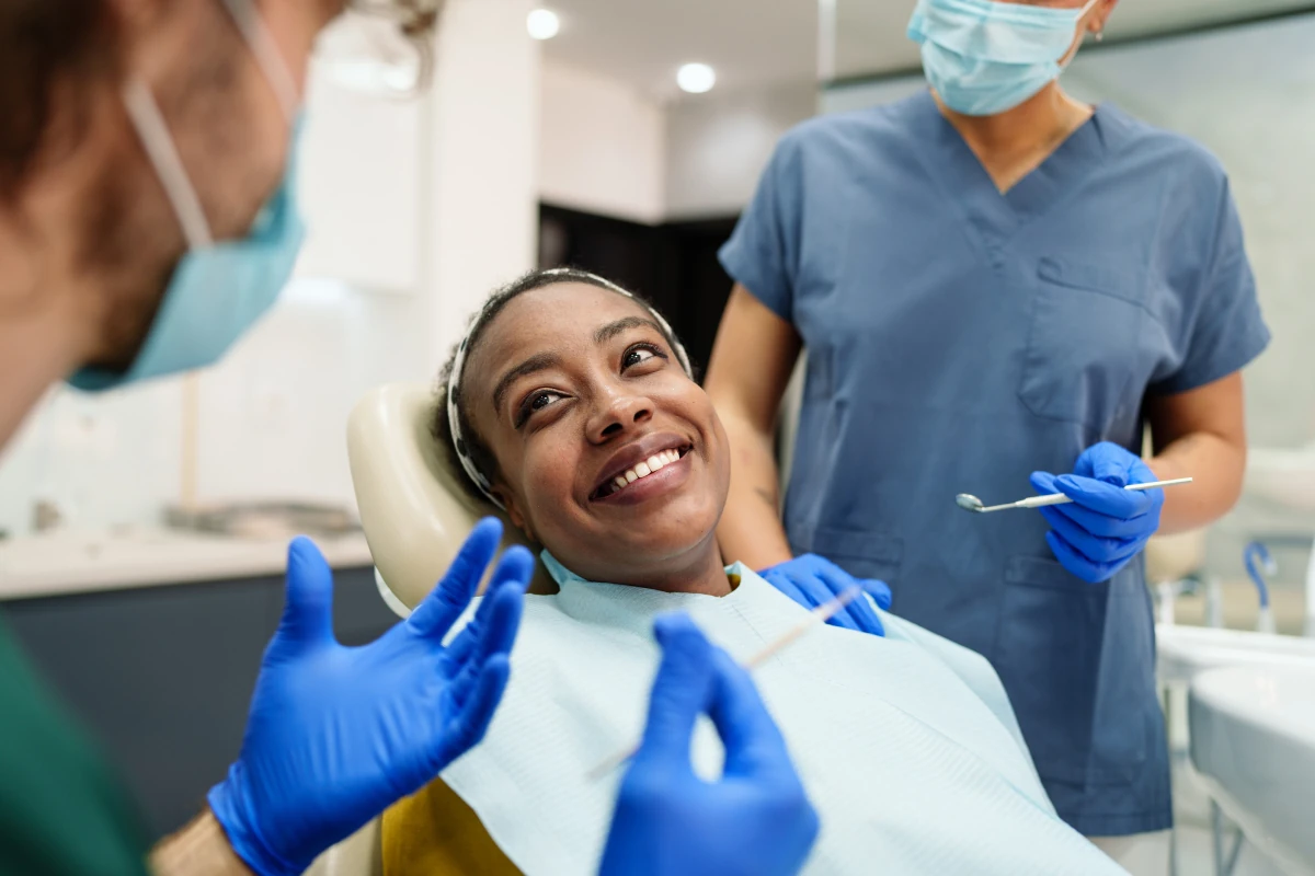 Smiling patient in a dental chair during a checkup with a dentist and assistant. Brightly lit modern clinic with professional medical tools.