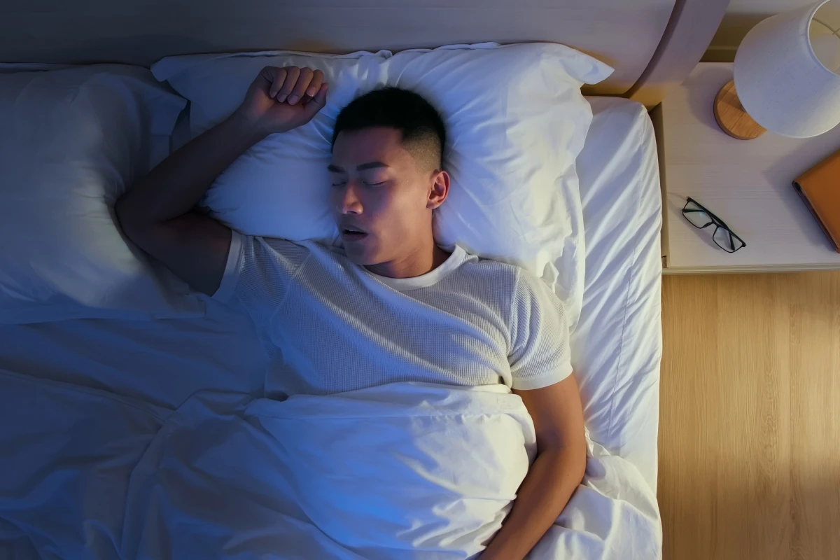 Man sleeping in white linens under cool blue ambient light and warm lamp glow. Top-down view of peaceful nighttime rest in a quiet bedroom.