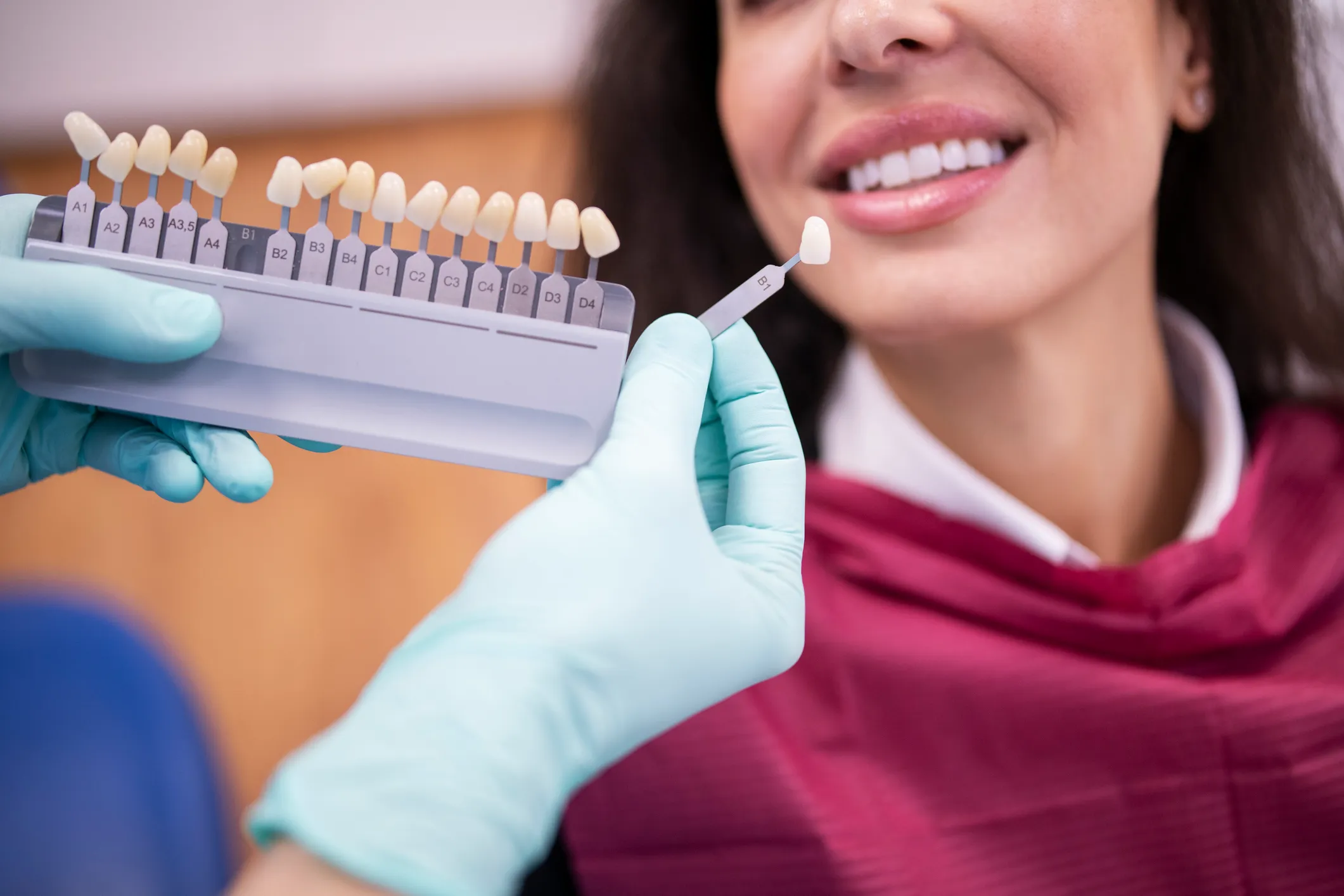 Dentist in blue gloves holds a VITA shade guide with A1-D4 tabs, color-matching a patient's teeth in a bright, professional clinic setting.