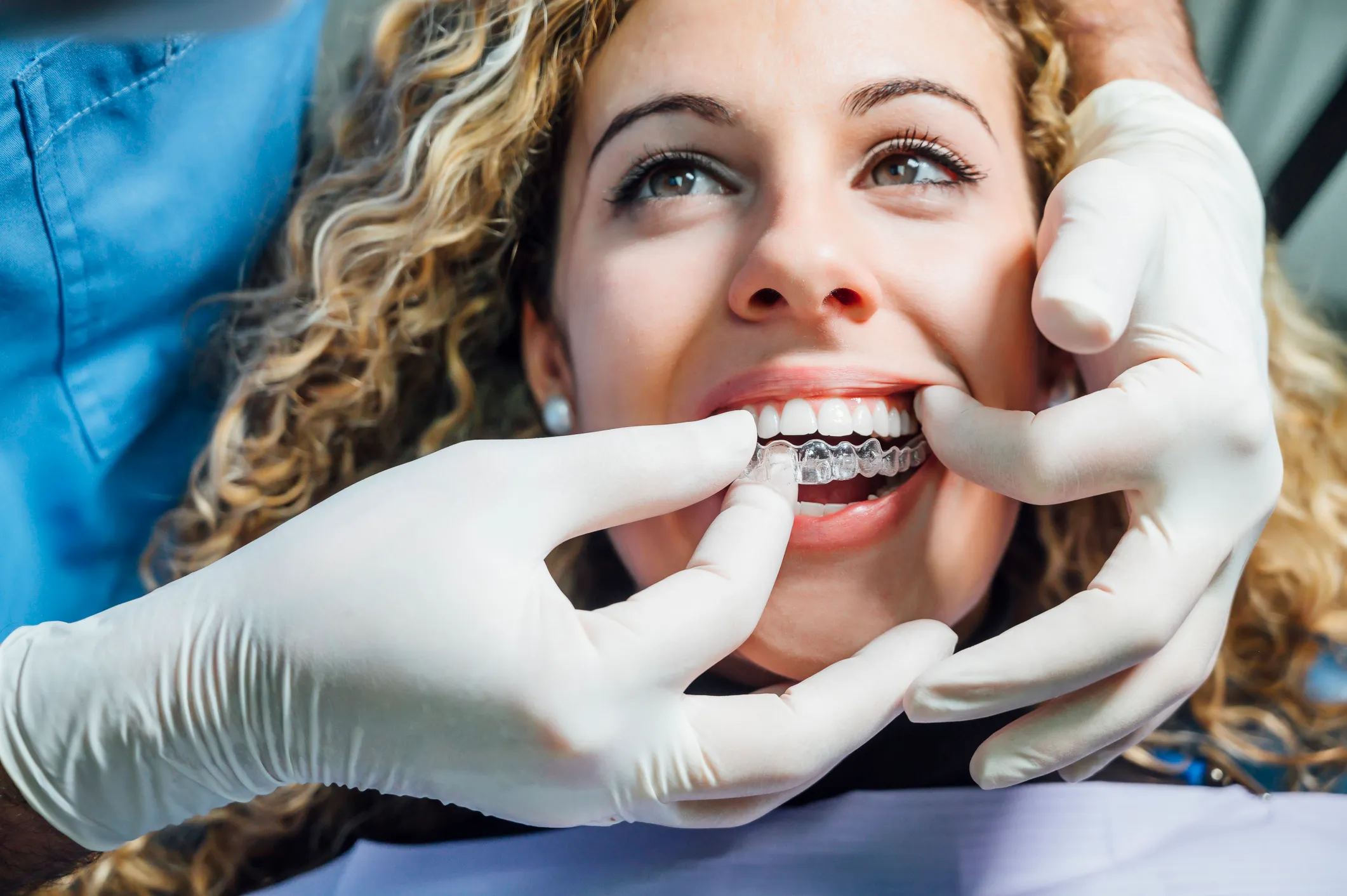 Dentist in blue scrubs and white gloves fitting a clear Invisalign aligner on a smiling patient under bright clinical dental office lighting.