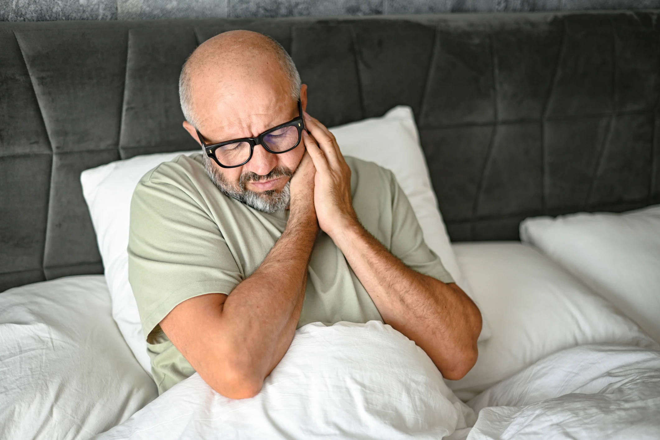 Bald man with glasses sitting in bed in soft bedroom lighting, clutching his jaw in pain with an expression of distress.